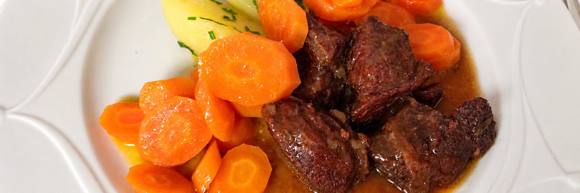 Gros plan sur une assiette blanche garnie de morceaux de bœuf mijoté en sauce, de rondelles de carottes et de pommes de terre à la ciboulette.