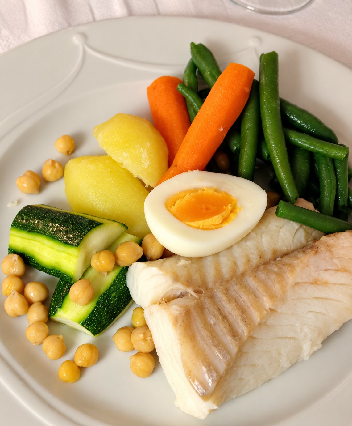 Une assiette blanche garnie de filet de poisson blanc, d'un demi-œuf dur, de haricots verts, de carottes, de pommes de terre, de courgette et de pois chiches.