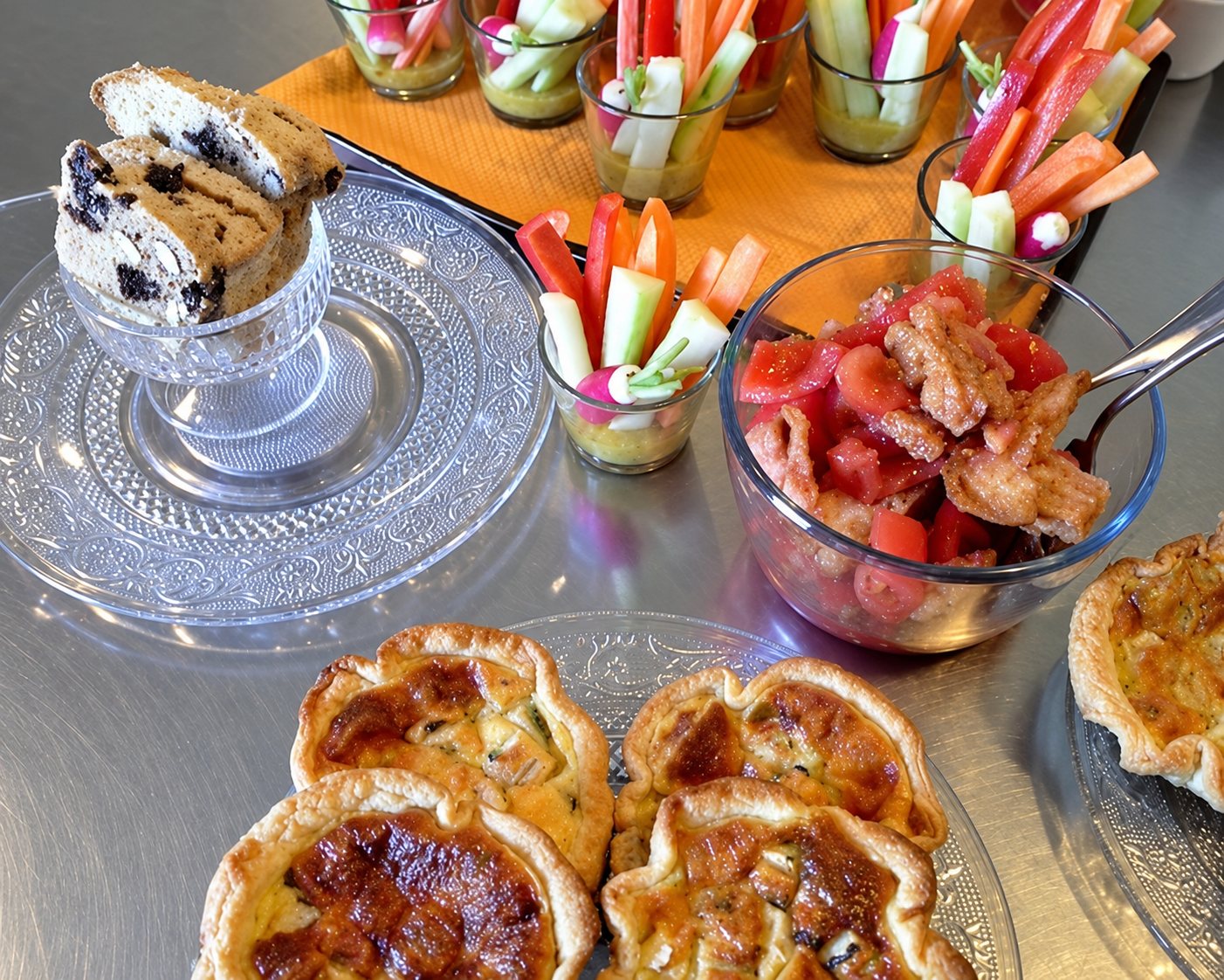 Table de buffet avec des tartelettes dorées, des verrines de bâtonnets de légumes crus, un saladier de tomates au jambon et des tranches de cake sur un support en verre.