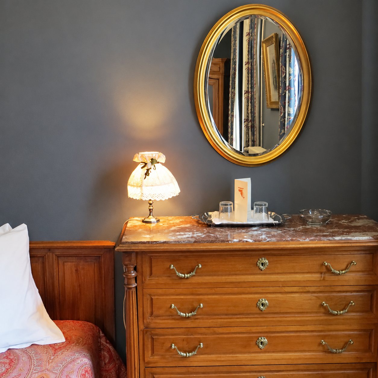 Coin de chambre avec une commode en bois à plateau de marbre, un miroir ovale doré, une petite lampe allumée et un plateau avec deux verres.
