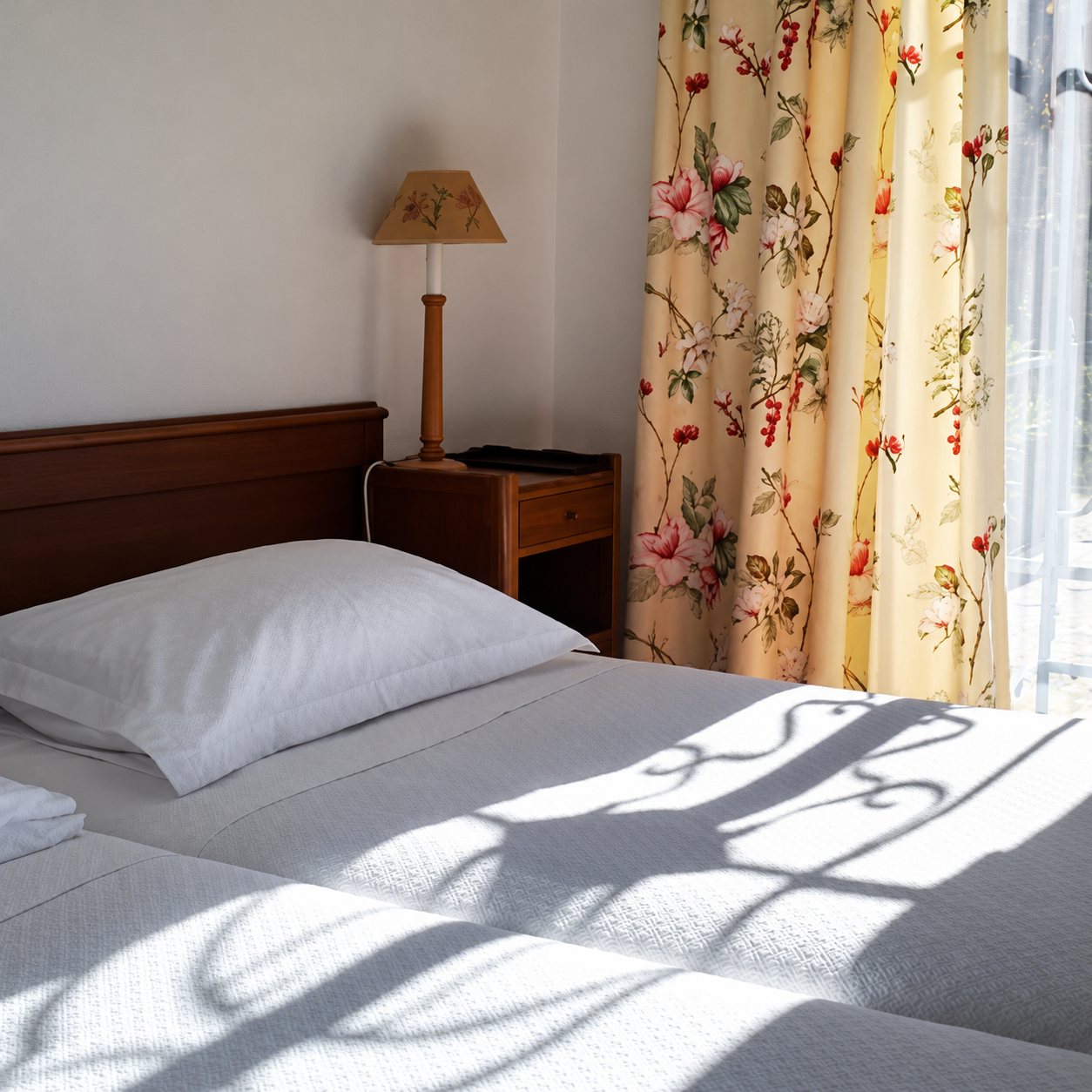 Chambre avec deux lits aux draps blancs, une lampe de chevet et des rideaux à motifs floraux, éclairée par le soleil qui projette l'ombre d'une grille en fer forgé.