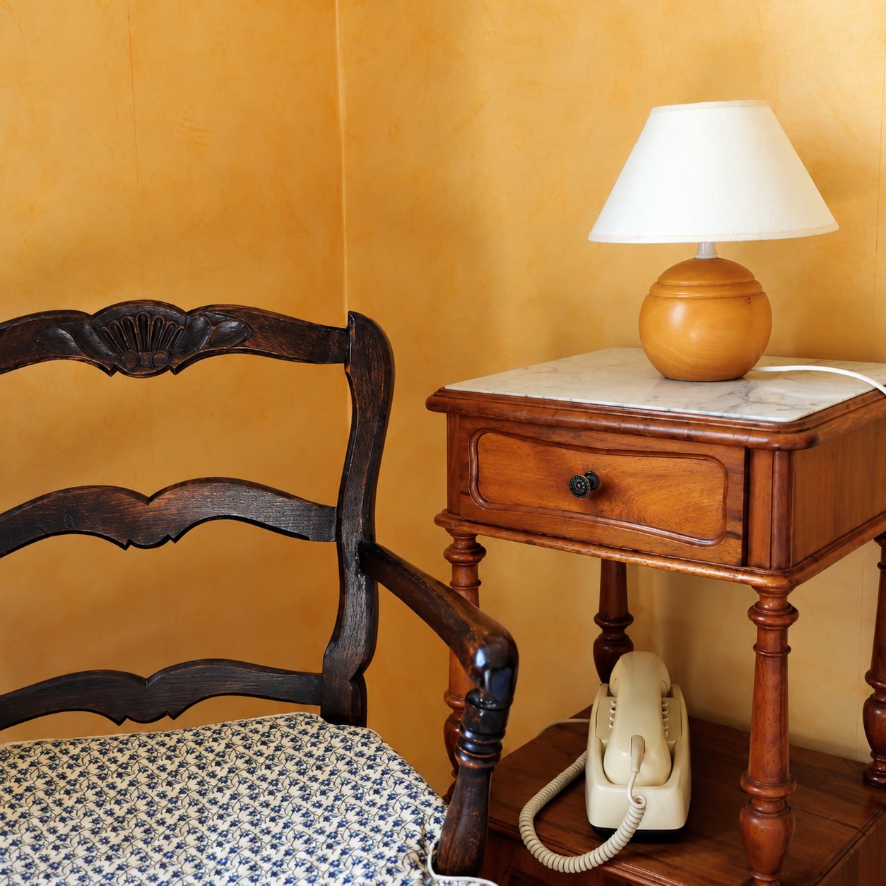 Un fauteuil en bois sculpté à l'assise fleurie est placé près d'une table de chevet ancienne à dessus de marbre, avec une lampe à abat-jour blanc et un téléphone beige, devant un mur ocre.