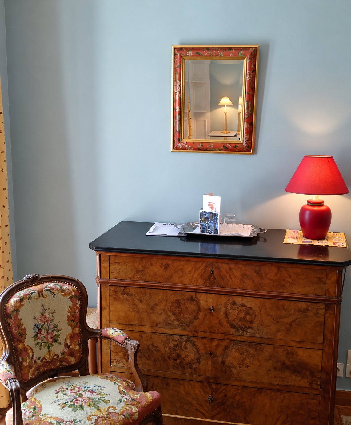 Dans une chambre aux murs bleu clair, une commode ancienne en bois à dessus noir porte une lampe rouge et un plateau, sous un miroir encadré, à côté d'un fauteuil de style à tapisserie fleurie.