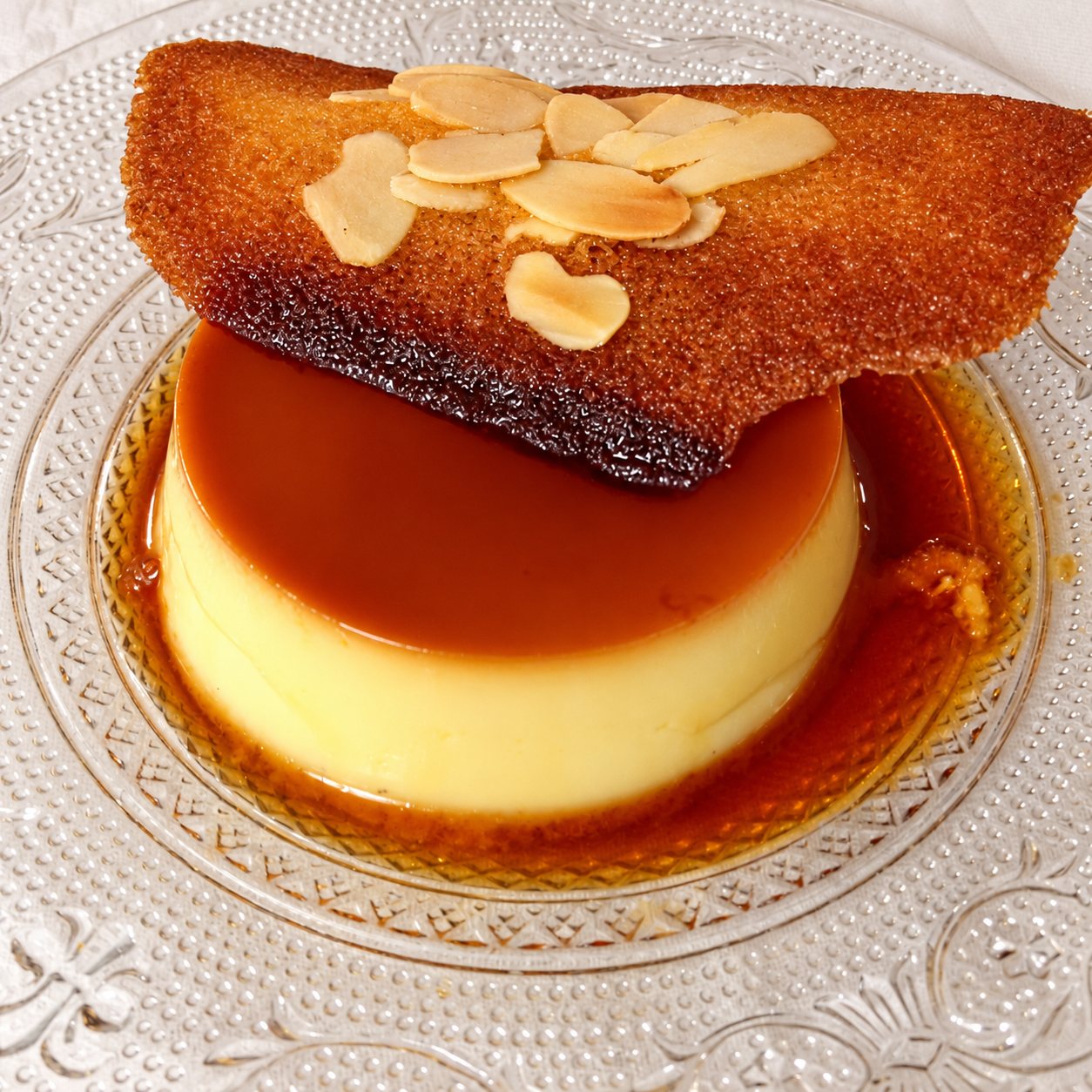 Une crème caramel nappée de caramel dans une assiette en verre ciselé, coiffée d'une fine tuile croustillante garnie d'amandes effilées.