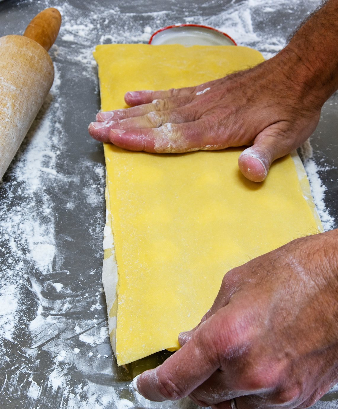 Deux mains couvertes de farine appuient sur une feuille de pâte jaune garnie, posée sur un plan de travail en inox à côté d'un rouleau à pâtisserie.