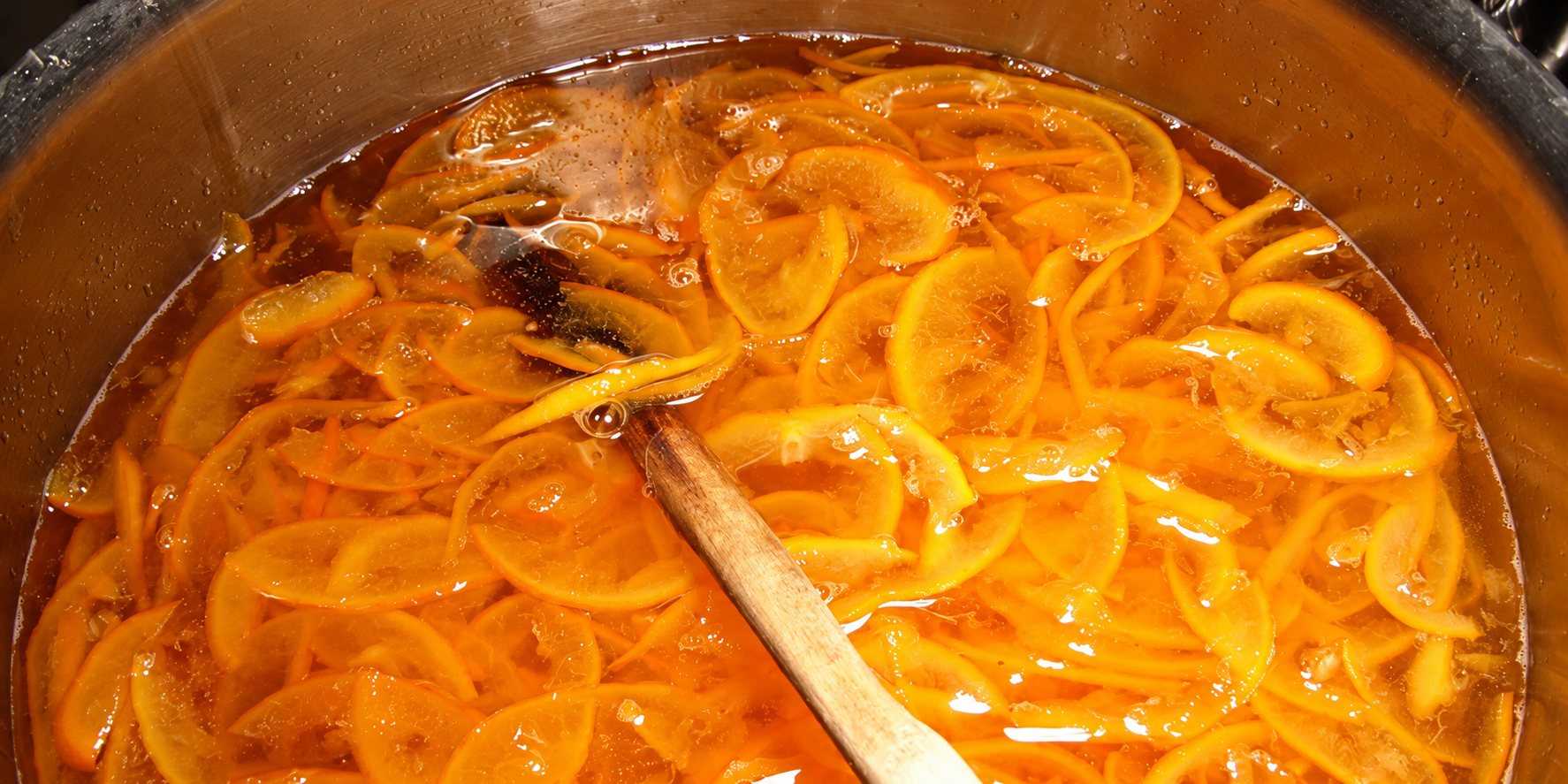 Des fines tranches d'orange mijotent dans un sirop doré au fond d'une grande marmite, avec une cuillère en bois qui plonge dans la préparation.