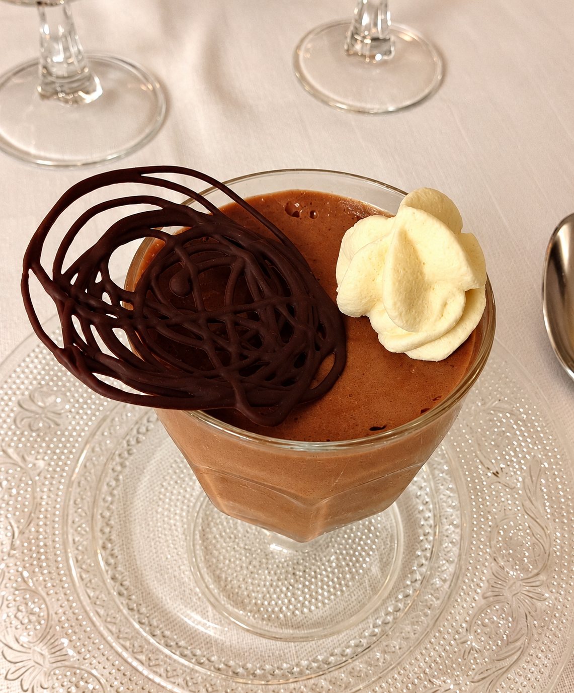 Une verrine de mousse au chocolat surmontée d'une dentelle de chocolat noir et d'une rosace de crème chantilly, posée sur une assiette en verre sur une nappe blanche.