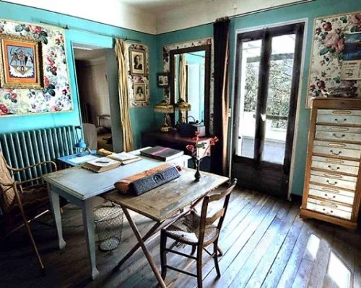 Intérieur lumineux aux murs turquoise décorés de tissus fleuris, avec une table de travail, des chaises en paille, un meuble à tiroirs et une porte-fenêtre donnant sur un balcon.