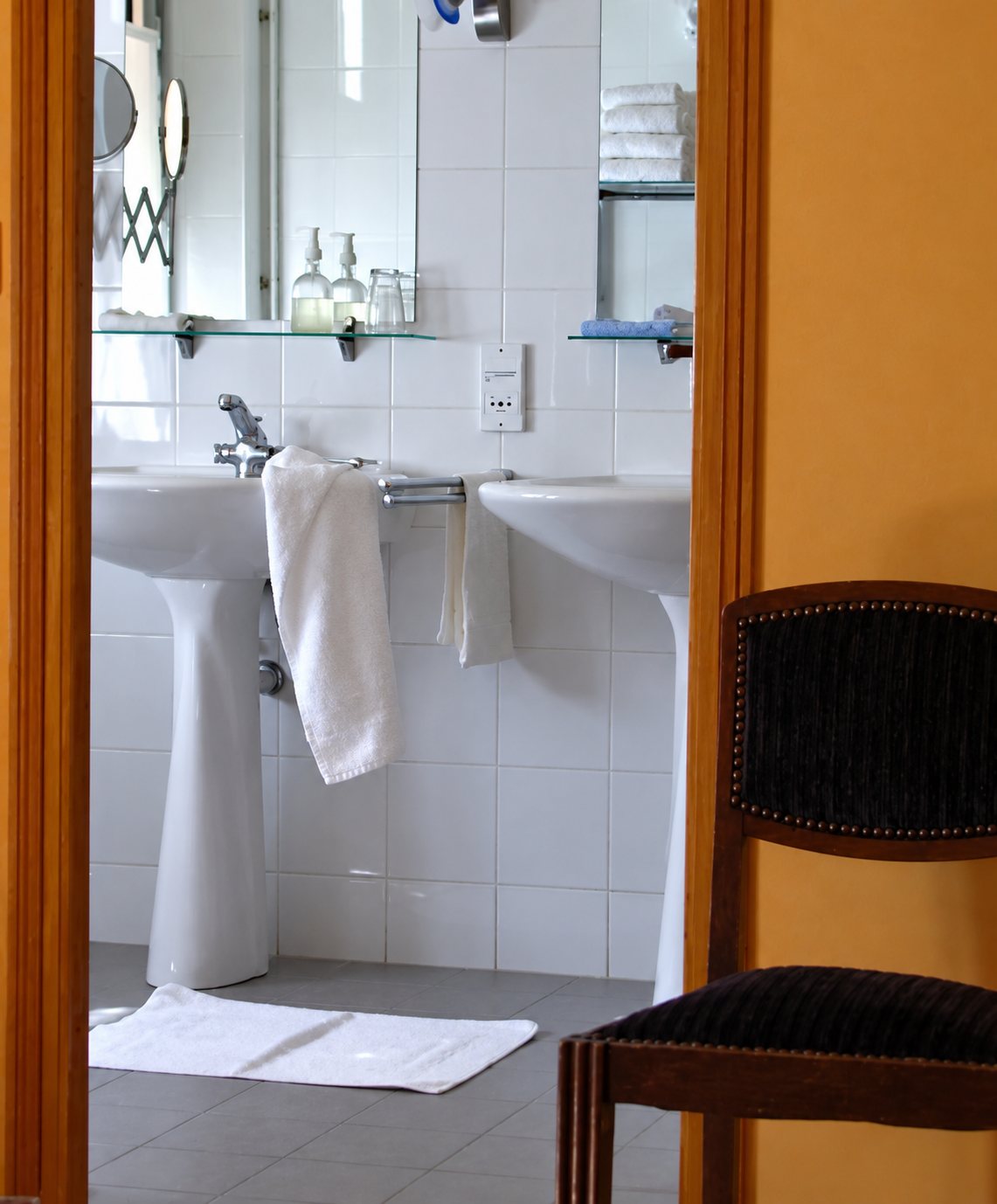 Vue à travers une porte d'une salle de bains aux murs carrelés de blanc, avec deux lavabos sur colonne, des serviettes blanches et une chaise au premier plan.
