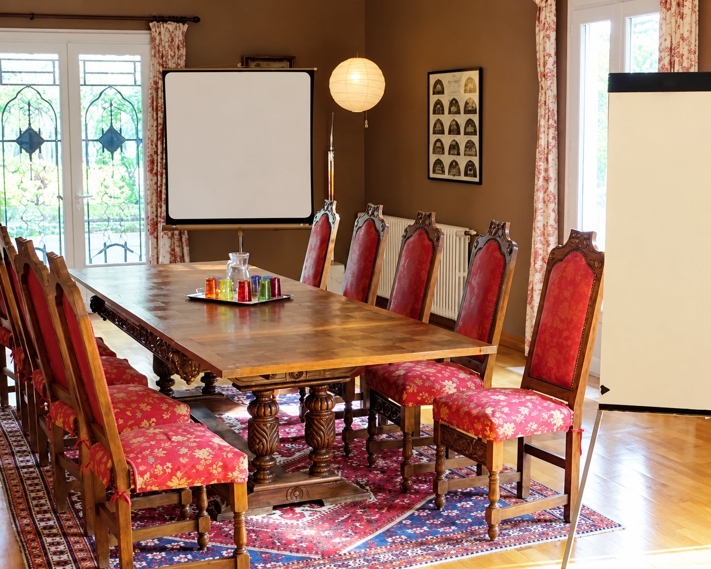 Une salle de réunion lumineuse avec une grande table en bois massif entourée de chaises à assise rouge, un écran de projection et un tapis coloré.