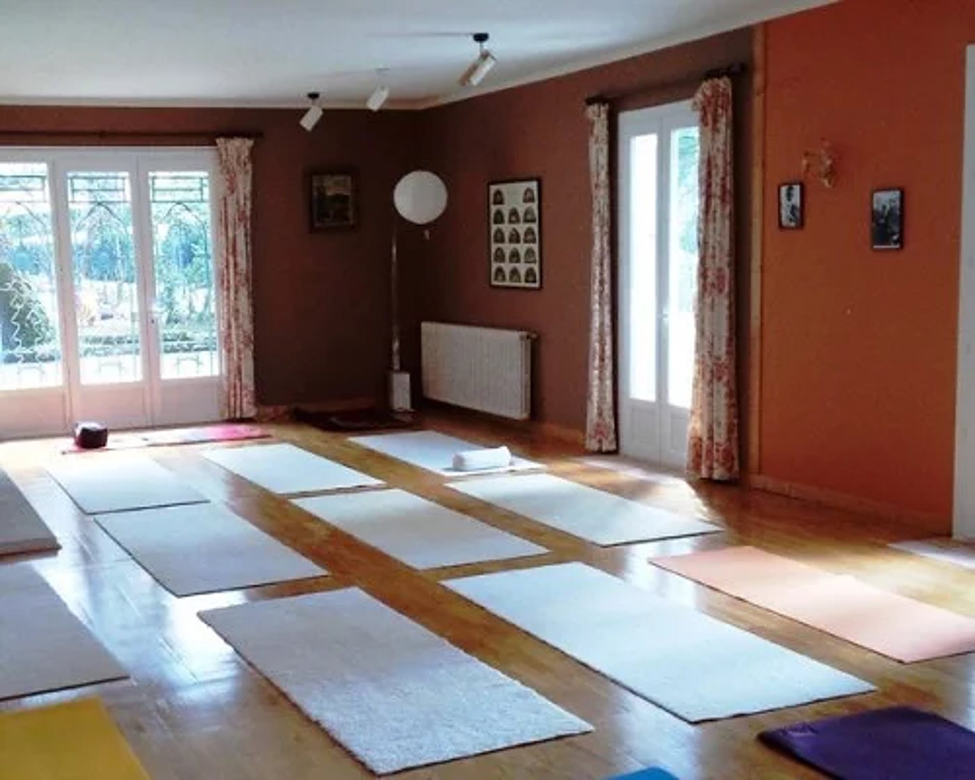 Une salle de yoga aux murs terracotta et au parquet ciré, avec plusieurs tapis blancs, roses et violets disposés au sol près de grandes portes-fenêtres.