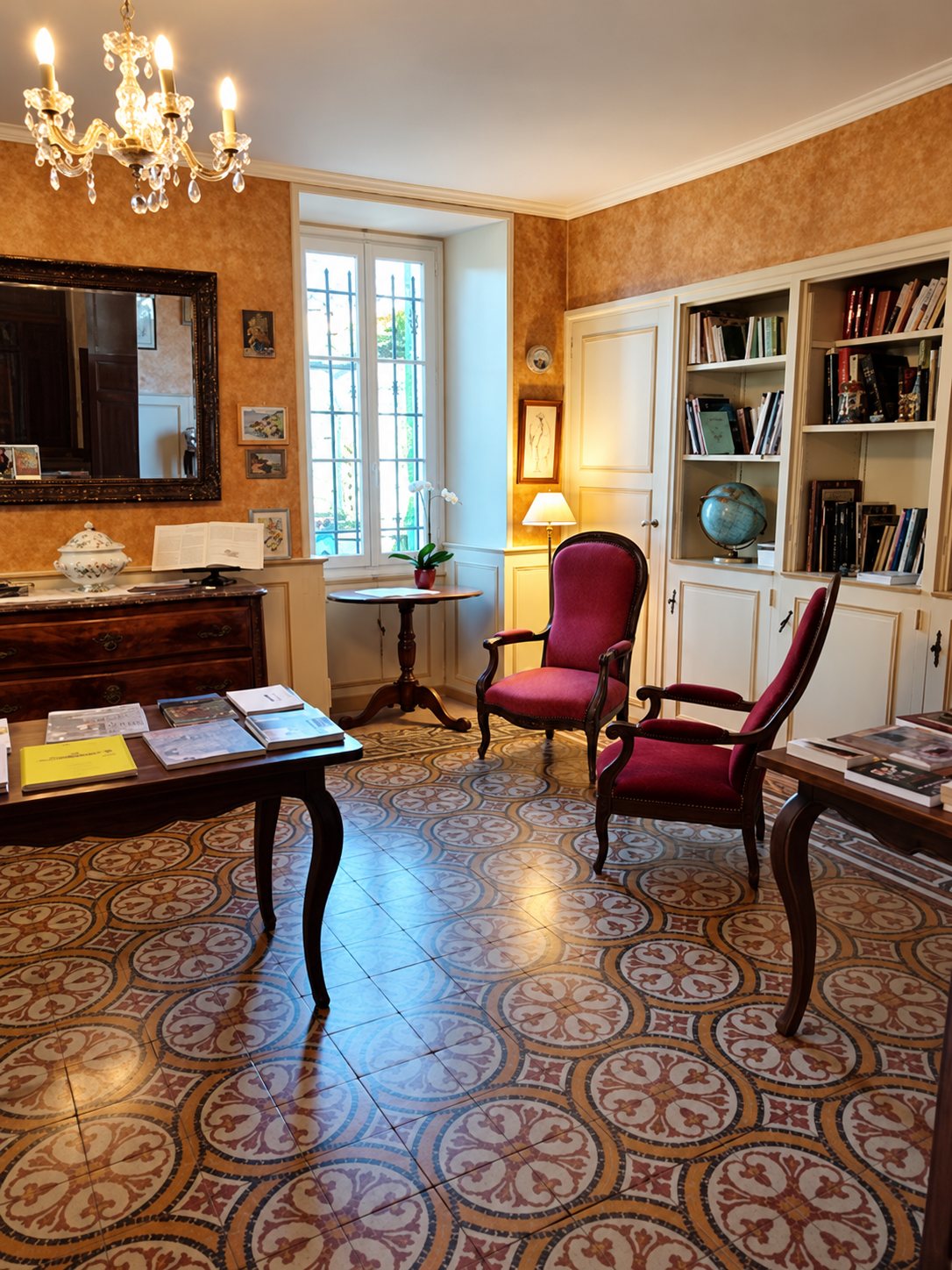 Un salon élégant avec deux fauteuils rouges, une bibliothèque blanche, un lustre et un sol en carreaux de ciment à motifs.