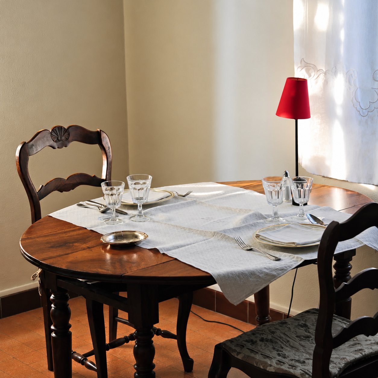 Une table ronde en bois sombre est dressée pour deux personnes avec verres, assiettes et couverts, à côté d'une lampe à abat-jour rouge et d'un rideau clair.