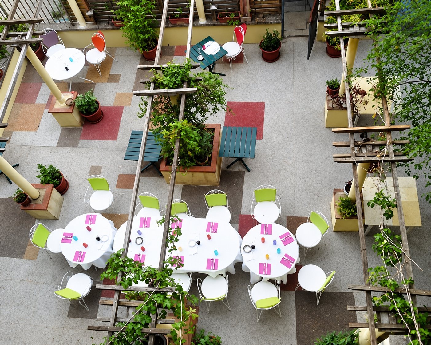 Vue du dessus d'une terrasse aménagée avec des tables rondes dressées, des chaises vertes et blanches, des plantes en pots et une pergola en bois.
